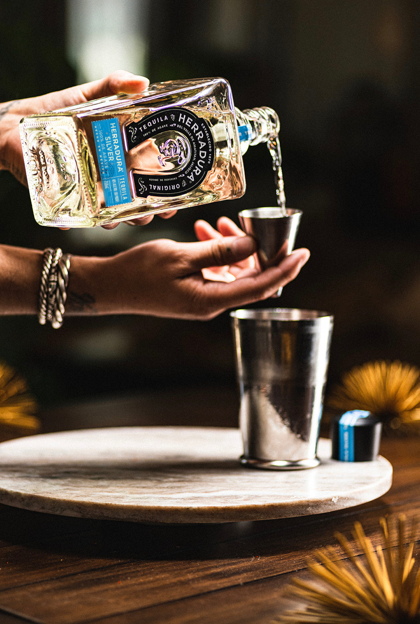A hand pours Herradura tequila into a glass
